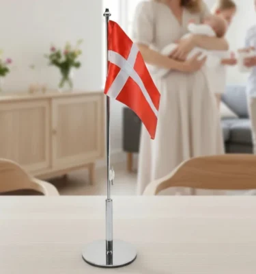 Et dansk bordflag på en metalstang stående på et lyst træbord med en sløret familie i baggrunden til en festlig begivenhed.